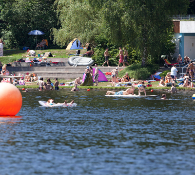 Das Strandbad vom Wasser aus. Es sind viele Menschen im Wasser und auf dem Strand.