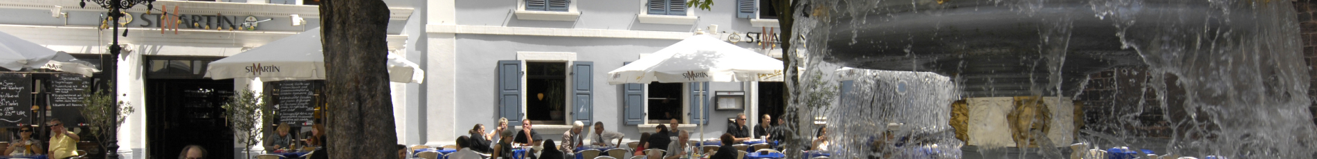 Der Martinsplatz im Sommer. Auf der Außenfläche vom Restaurant St. Martin sitzen bereits einige Leute. Rechts im Bild ist der Martinsbrunnen mit seinen Schwänen zu sehen.