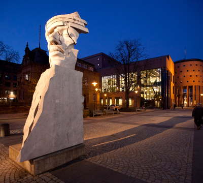 Das Pfalztheater bei Dämmerung. Davor die aus Stein gemeißelte Schauspiel Maske.