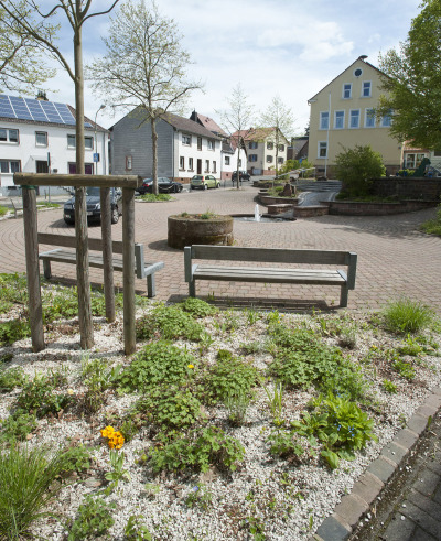 Ein gepflasterter Platz in Dansenberg mit Blick auf die Dansenberger Straße. Auf dem Platz befindet sich ein kleiner, im Boden versunkener, Brunnen und mehrere Bänke als Sitzgelegenheit.