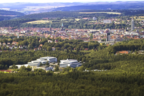 Eine Ansicht über die Stadt vom Humbergturm aus. Rund um Kaiserslautern liegt der grüne Pfälzer Wald und im Stadtkern ragen mehrere Kirchentürme hoch hinauf. Auch das Rathaus ragt aus der Häusermenge, die immer wieder von Wald abgewechselt wird.