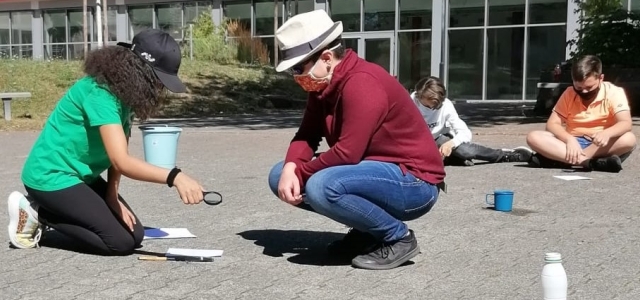 Zwei Kinder sitzen auf dem Steinboden vor einem Schulgebäude. Eines der Kinder hält eine Lupe in der Hand. Beide tragen Kopfbedeckungen und Gesichtsmasken.