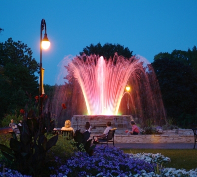 Parkbesucher in der Dämmerung sitzen um einen Brunnen mit illuminierter Wasserfontäne