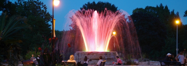 Parkbesucher in der Dämmerung sitzen um einen Brunnen mit illuminierter Wasserfontäne