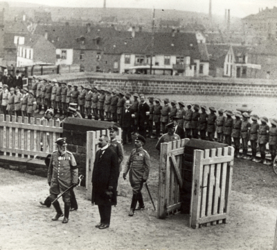 König Ludwig III. trägt Militäruniform und Degen. Er steht neben Oberbürgermeister Küfer, die einzige Person in Zivil. Hinter ihnen versammeln sich in zwei Reihen Soldaten.